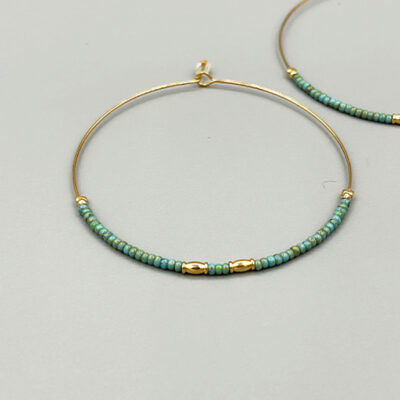 Detail boucles créoles larges dorées à l'or 18 carats avec perles de verre japonaises turquoises. Packshot sur fond gris.