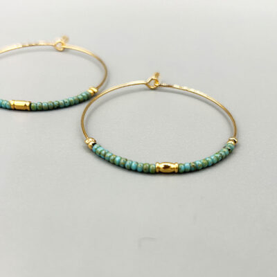 Detail boucles créoles taille moyenne dorées à l'or 18 carats avec perles de verre japonaises turquoises. Packshot sur fond gris.