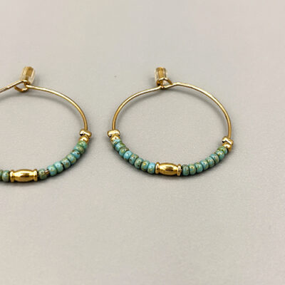 Detail boucles créoles petites dorées à l'or 18 carats avec perles de verre japonaises turquoises. Packshot sur fond gris.