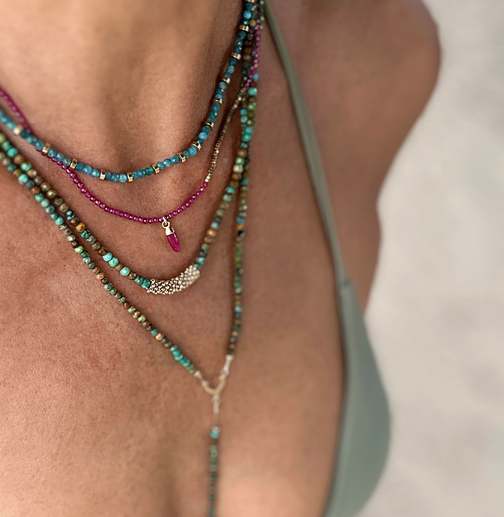 Gros plan sur un décolleté orné de plusieurs colliers fins superposés aux perles turquoise, dorées et fuchsia, avec un pendentif en forme de goutte.