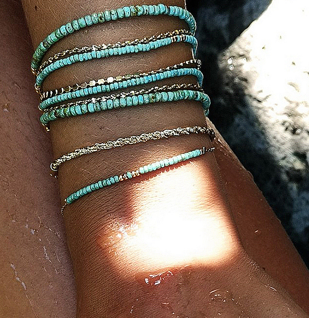 Gros plan sur un poignet paré de plusieurs bracelets fins en perles turquoise et dorées, empilés en accumulation, avec un reflet de soleil sur la peau bronzée.